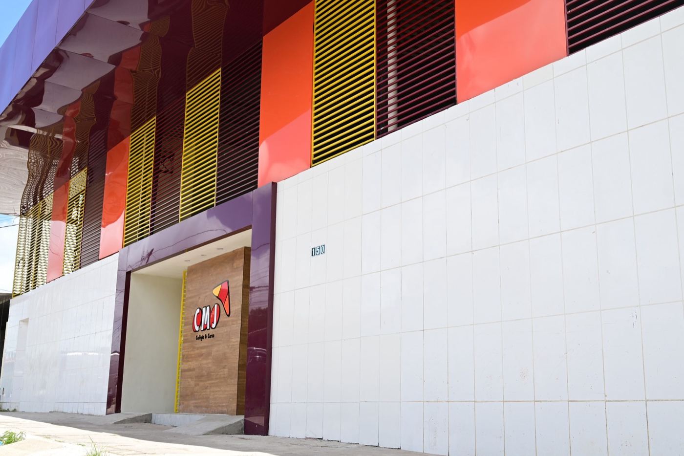 Fachada do CMJ com brises amarelos, laranja e vinho e a entrada com o logo do colégio