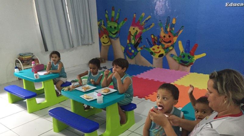 Crianças pequenas lanchando em mesas coloridas com a professora