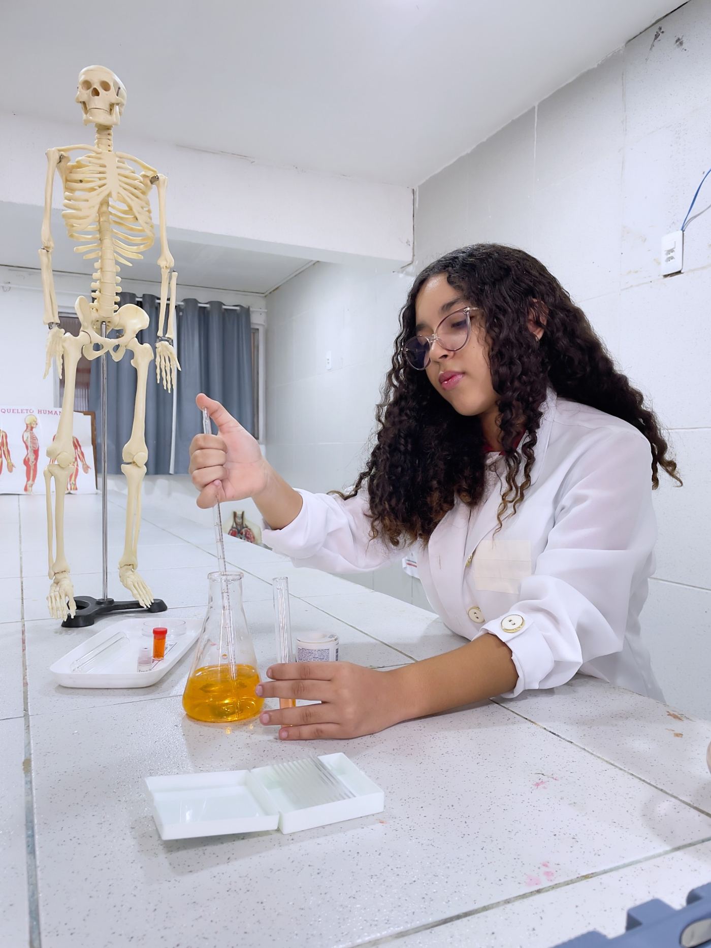 Estudante de jaleco fazendo experiência no laboratório de ciências, ao lado de um esqueleto humano