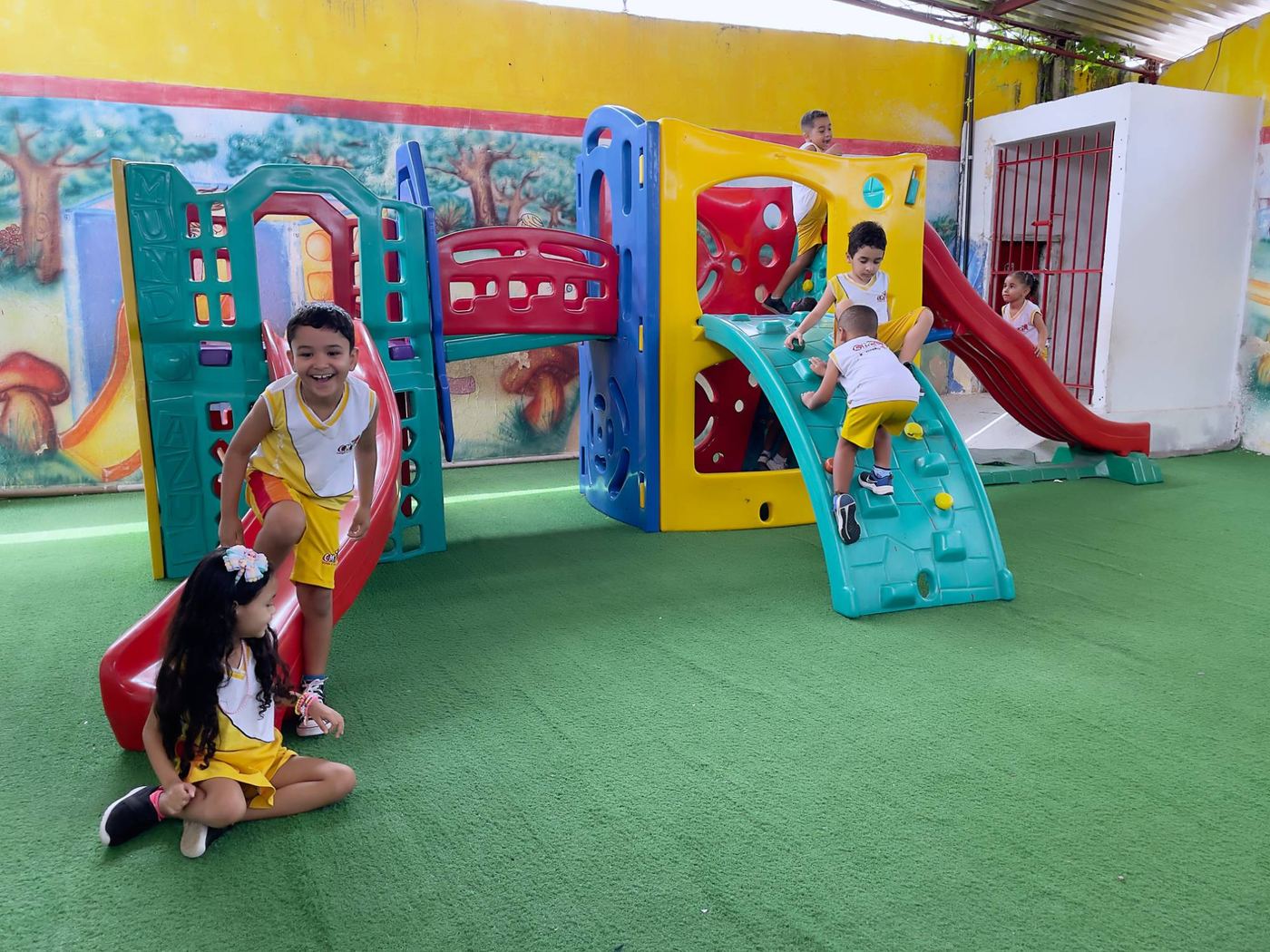 Crianças da Educação Infantil brincando no parquinho colorido, com escorregadores, sobre grama sintética