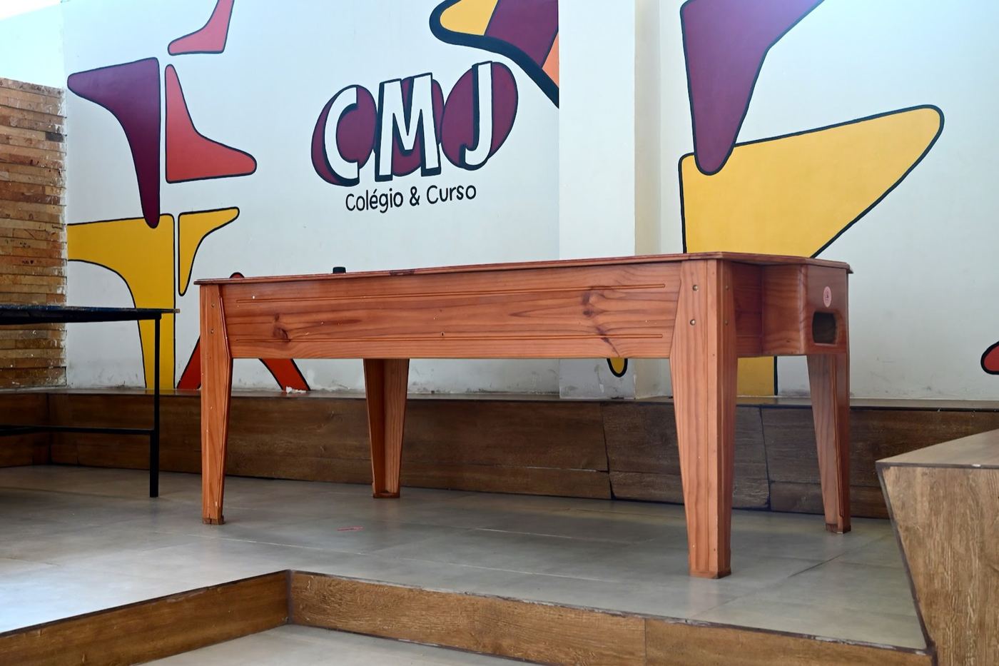Mesa de pebolim de madeira diante de um mural com o logo do CMJ