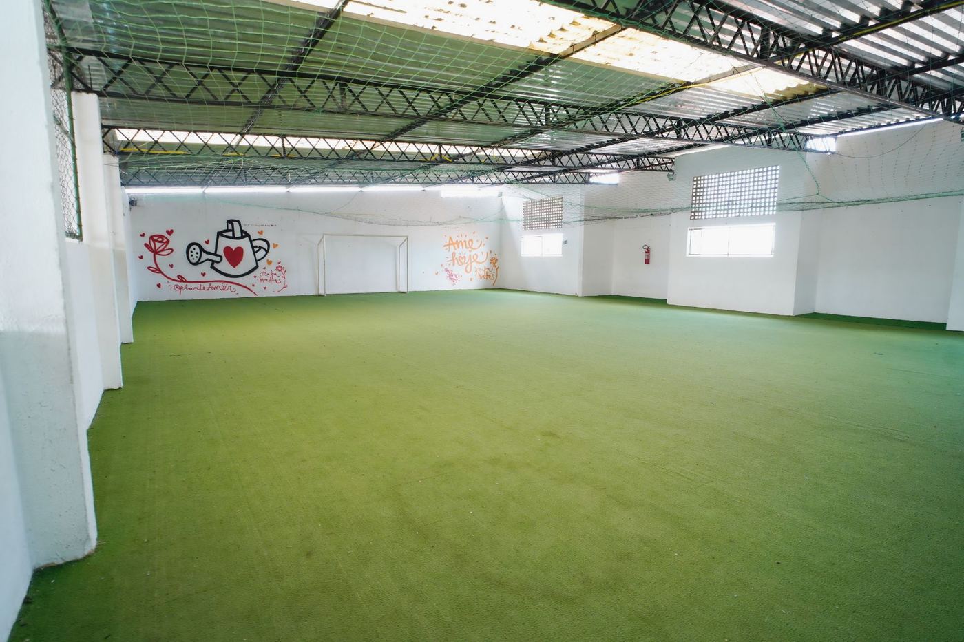 Quadra coberta com piso de grama sintética, trave de futebol e grafites nas paredes brancas