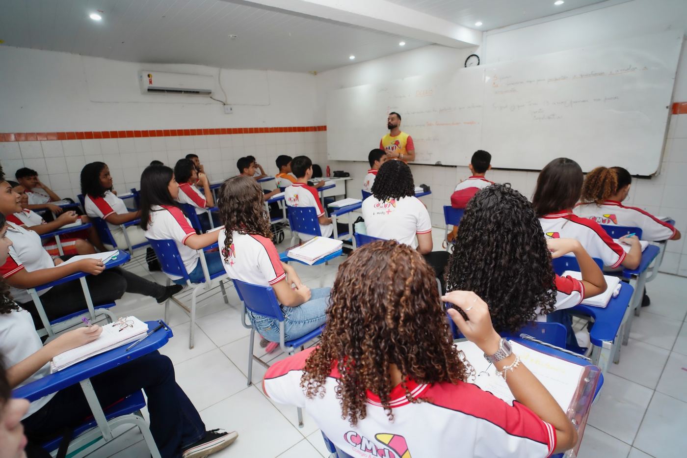 Turma do Fundamental II do CMJ de uniforme em sala climatizada, com o professor no quadro