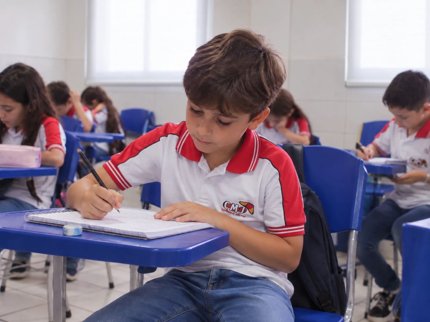 Estudante do Fundamental I do CMJ, de uniforme, escrevendo no caderno em sala de aula com os colegas ao fundo